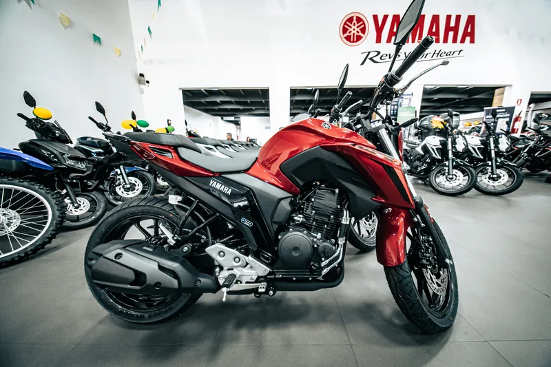 Fazer FZ25 vermelha vista de lado, com a parede Yamaha ao fundo.