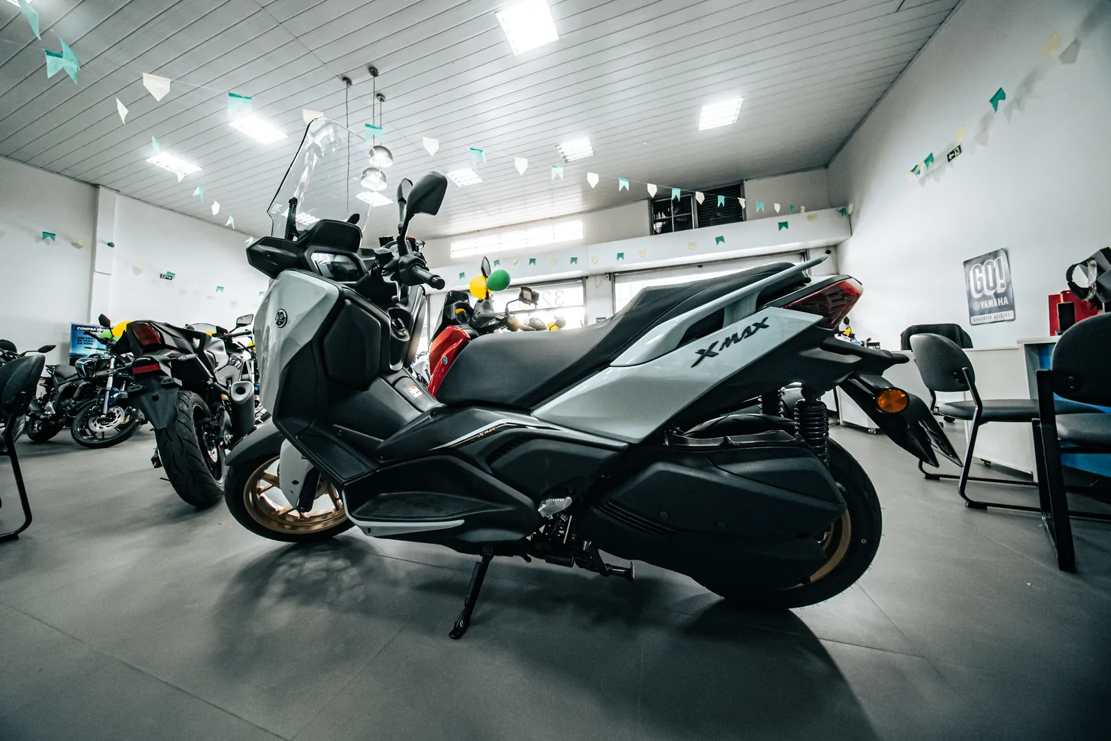 XMAX 250 cinza com rodas douradas, vista de lado no salão da loja.